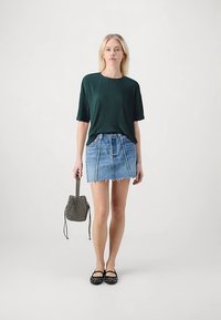 Modelo vistiendo una camiseta de manga corta verde oscuro acanalada, minifalda vaquera con dobladillo deshilachado, flats negros con tachuelas y un bolso tipo bucket con estampado.