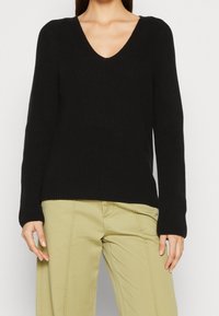 Frau in einem schwarzen V-Ausschnitt Langarmpullover und hellbeigen Hosen, stehend vor einem einfarbigen weißen Hintergrund.