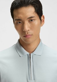 Ljusblå polo shirt med dragkedja i kragen, med en strukturerad tygkvalitet och en kontrasterande mörk linje längs dragkedjan. Närbild.