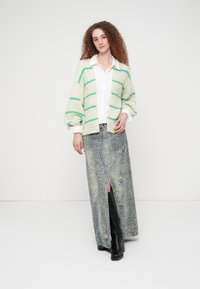 Πλεκτή ζακέτα με πράσινες ρίγες πάνω από λευκό πουκάμισο, συνδυασμένη με φούστα γκρι με σχέδιο φίδι και μπροστινή σχισμή, καθώς και μαύρα μπότες μέχρι το γόνατο.