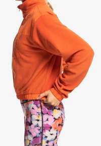 Giacca in fleece arancione con colletto alto, tasca frontale e polsini elasticizzati; abbinata a leggings con motivo floreale multicolore.
