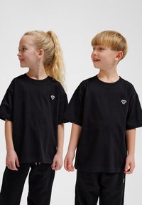 To børn iført løstsiddende sorte bomulds t-shirts med korte ærmer, der har et lille hvidt logo på venstre side af brystet.