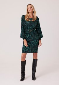 Kobieta w sukience w zielono-czarny wzór zebry z paskiem, długimi rękawami, złotym pierścionkiem i czarnych kozakach na wysokim obcasie do kolan, stojąca na jednolitym tle.