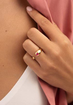 Anillo de oro con una piedra preciosa rosa en forma de pera y un pequeño acento de perla de oro, exhibido en una mano con un fondo de tela rosa.
