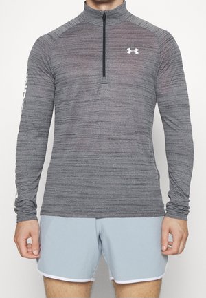 Mann trägt langärmeliges graues Sportoberteil mit Viertelreißverschluss und Under Armour Logo sowie hellblaue Shorts mit weißem Rand, steht vor einfarbigem Hintergrund.