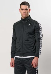 Mężczyzna w czarnej kurtce sportowej na zamek i spodniach z białymi paskami z logo na rękawach i nogawkach, stojący na jednolitym tle.