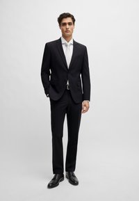 Fekete szabott öltöny, fehér inggel és világos nyakkendővel. Fekete bőr cipők, slim fit. Simább textúra, klasszikus hajtókák.