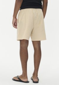 Vyras stovi, vilkėdamas smėlio ir baltas juostuotas šortus, juodą berankovį marškinėlį ir juodus šlepetes, pavaizduotas iš nugaros prieš baltą foną.