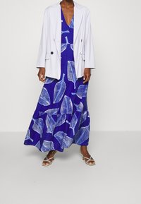 Blå mönstrad maxiklänning med vita bladmotiv, kombinerad med en vit blazer och sandaler med remmar. Slätt tyg, avslappnad passform och lagerdämpad nederdel.