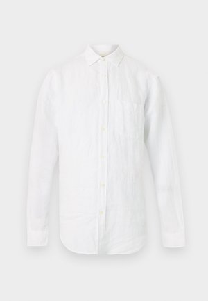 Mann mit kurzen blonden Haaren, trägt ein weißes Hemd mit Knöpfen, blaue knielange Jeansshorts und schwarze elegante Schuhe, steht mit den Händen in den Taschen.