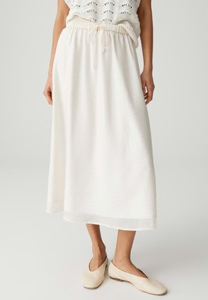 Frau trägt einen weißen Midirock mit Kordelzug in der Taille, ein beigefarbenes Strickoberteil und cremefarbene flache Schuhe und steht vor hellem Hintergrund.