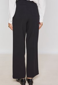 Zwarte wide-leg broek met een gladde textuur, voorzien van twee achterzakken en een aansluitende tailleband, gedragen met glanzende zwarte schoenen.