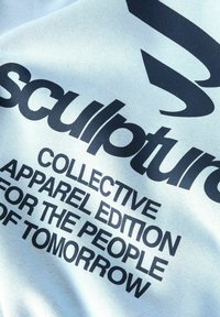Ljusblått tyg med svart text som står "skulptur KOLLEKTIV KLÄDER EDITION FÖR MÄNNISKORNA IMORGON."