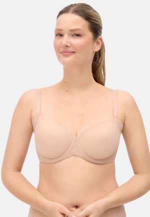 Mujer de piel clara y cabello castaño recogido en una coleta, lleva un sujetador acolchado de color nude, de pie frente a un fondo claro y sencillo.