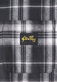Black and gray plaid fabric with a soft texture. Features a yellow "Stun Ray" logo on a black label. Includes a chest pocket.