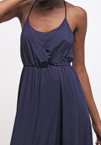 Marineblauwe, mouwloze jurk van zacht, vloeiend materiaal. Heeft een wikkelontwerp bij het lijfje en een elastische tailleband voor een goede pasvorm.