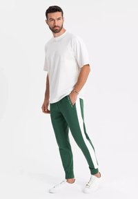 Vit bomullströja med korta ärmar, parat med gröna joggers med vita sidostrikar, och vita sneakers. Modellen står med händerna i fickorna.