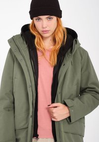 Veste extérieure vert olive avec une fermeture éclair et des boutons, superposée sur un hoodie noir et une chemise rose clair. Comprend des poches et une capuche doublée.