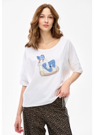 Kobieta o falowanych brązowych włosach, ubrana w białą koszulę z nadrukiem czytelniczym oraz spodnie w panterkę z bocznymi lampasami.