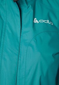 Tyrkysová bunda Odlo s hladkou strukturou, zipovým uzávěrem a logem na hrudi, s jemnými geometrickými vzory v látce.