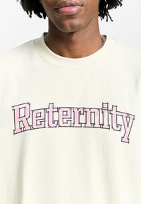 Kremfarget hettegenser med en stor, rosa trykt "Reternity"-logo. Myk stoff med rund hals og avslappet passform.