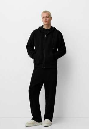 Schwarze Zip-Hoodie mit Kängurutasche, kombiniert mit passenden schwarzen Hosen. Glatte Textur, lockere Passform, vervollständigt mit weißen Sneakern.