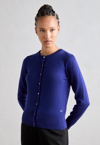 Royal blue kardigán, könnyű anyagból készült, körkivágással, hosszú ujjakkal és elülső gombokkal. A szélei bordás díszítéssel vannak ellátva.