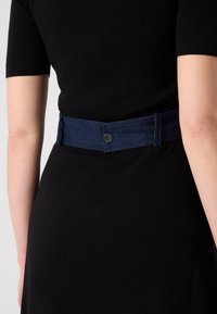 Ceinture en denim bleu foncé avec une texture lisse, dotée d'une fermeture à pression centrale et d'un design large. Portée sur une robe noire.