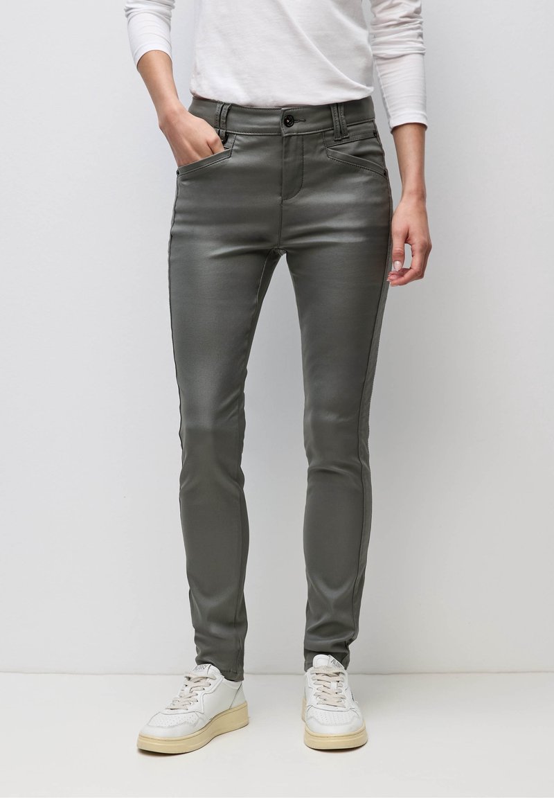 Personne portant un pantalon gris foncé ajusté, un haut blanc à manches longues et des baskets blanches, debout devant un fond uni.