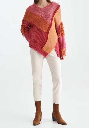 Woman wearing color-blocked pink and orange knit sweater with tassels, white pants, and brown suede ankle boots.