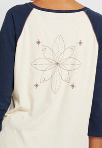 Krämfärgad skjorta med marinblå trekvartslånga ärmar; har en broderad blomsterdesign i burgund på baksidan. Mjuk textilfinish.