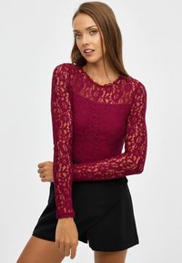 Top de encaje burdeos de manga larga con patrón floral y escote con puntilla, combinado con pantalones cortos negros. Textura transparente visible en las mangas.