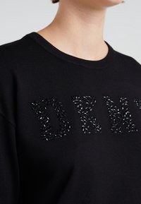 Svart stickad sweatshirt med bokstäverna "DRNY" prydda med glittrande svarta och grå pärldetaljer. Rund halsringning.
