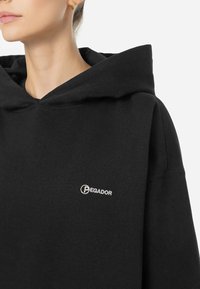 Svart hoodie i mjukt material, med en stor huva och broderad "Pegador"-logotyp i vitt på bröstet. Enkel och avslappnad passform.