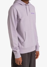 Persona che indossa un hoodie viola chiaro con tasca frontale e logo "ELEMENT" sul petto, abbinato a pantaloni scuri, su uno sfondo bianco.