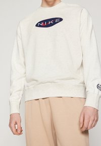Krämfärgad sweatshirt i mjukt tyg med rund hals, prydd med en marinblå och röd broderad Nike-logotyp på bröstet och ribbade muddar.