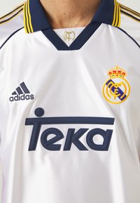 Maillot blanc avec un col marine, orné de rayures jaunes. Affiche "Teka" en marine et un logo rond avec une couronne et un emblème sur la poitrine.