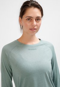 Hellgrünes, langärmliges Sportoberteil aus strukturiertem Stoff. Mit Rundhalsausschnitt und einem auf der Brust aufgedruckten Logo.