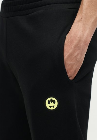 Χέρι τοποθετημένο στην τσέπη μαύρου παντελονιού με κίτρινο κυκλικό σήμα που παρουσιάζει τρία μαύρα αποτυπώματα πατούσας και βρυκολακίσια δόντια.