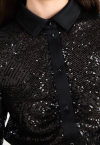 Close-up van een zwart met pailletten versierd shirt met een zwarte kraag en een verticale knoopsluiting met vierkante zwarte knopen.