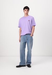 Purple cotton t-shirt with a small logo, paired with light blue, loose-fit jeans. Model wears black shoes, stands against a plain background.