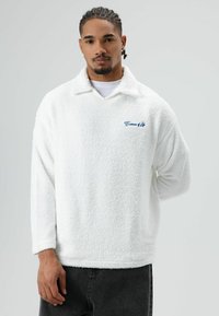 Witte, pluizige trui met een kraag en lange mouwen. Voorzien van blauwe borduursels op de borst. Het materiaal oogt zacht en heeft een textuur.