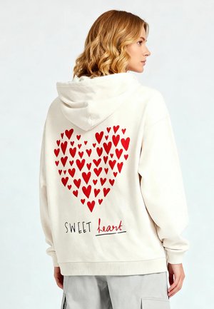 Crèmekleurige hoodie met een hartgrafiek gemaakt van rode harten op de achterkant, met de tekst "SWEET heart" onder het ontwerp. Zachte stof, ontspannen pasvorm.