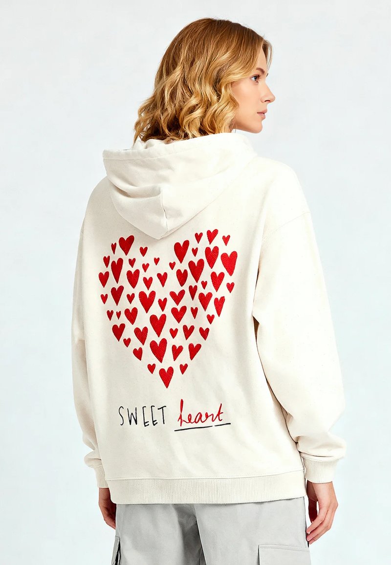 Felpa crema con una grafica a forma di cuore composta da cuori rossi sul retro, con il testo "SWEET heart" sotto il design. Tessuto morbido, vestibilità rilassata.