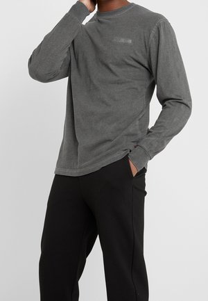 Personne portant un t-shirt à manches longues gris foncé délavé avec le texte "Copenhagen" et un pantalon noir, main dans la poche, sur un fond clair uni.