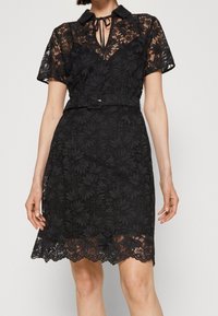 Robe en dentelle noire à manches courtes, col et fermeture à nouer. Motif floral, taille ceinturée, ourlet festonné. Texture en superposition transparente.