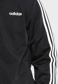 Giacca nera adidas con accenti bianchi a tre strisce sulle maniche, texture liscia e piccolo logo sul petto.