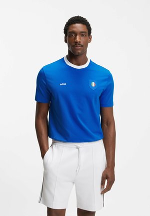 Homme portant un T-shirt bleu à col rond avec un col blanc et un emblème, assorti à un short blanc à cordon de serrage, se tenant avec une main dans la poche.