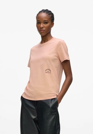 Femme aux cheveux tressés portant un T-shirt rose clair Karl Lagerfeld et un pantalon en cuir noir, mains dans les poches, regardant devant elle.