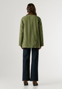 Olīvu zaļa jaka ar brūnu apkakli, taisna griezuma, ar spiedpogu aizdari sānos, apvienota ar tumšiem denima plaša griezuma biksēm.